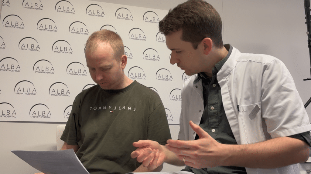 To mænd i en konsultation, den ene iført en Tommy Jeans t-shirt, gennemgår dokumenter i et rum med Alba Hårtab logoer.
