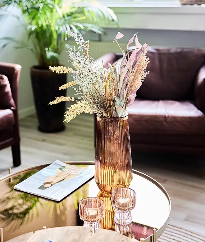 Vase med tørrede blomster og to glas på et bord, med en magasin i baggrunden.