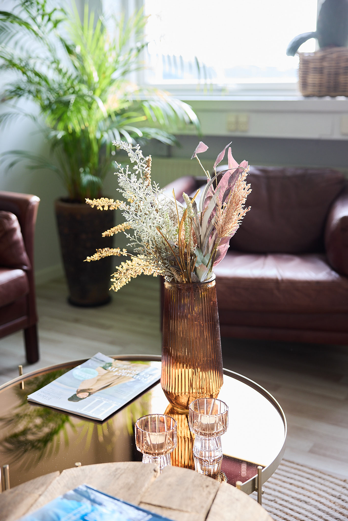 Vase i brunlig farve med tørrede blomster, placeret på et bord med et magasin og to glas.
