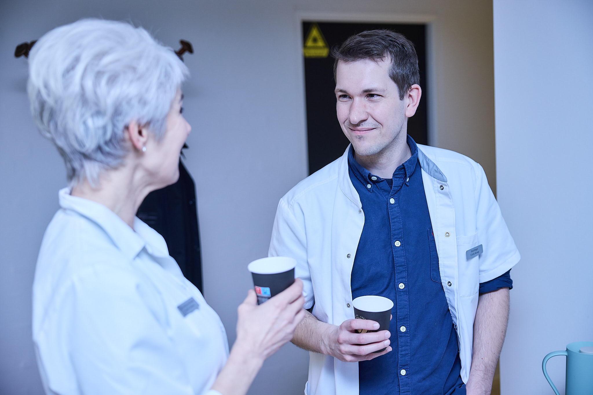 To personer i hvide labcoats, der holder papkopper og taler sammen i et klinisk miljø.