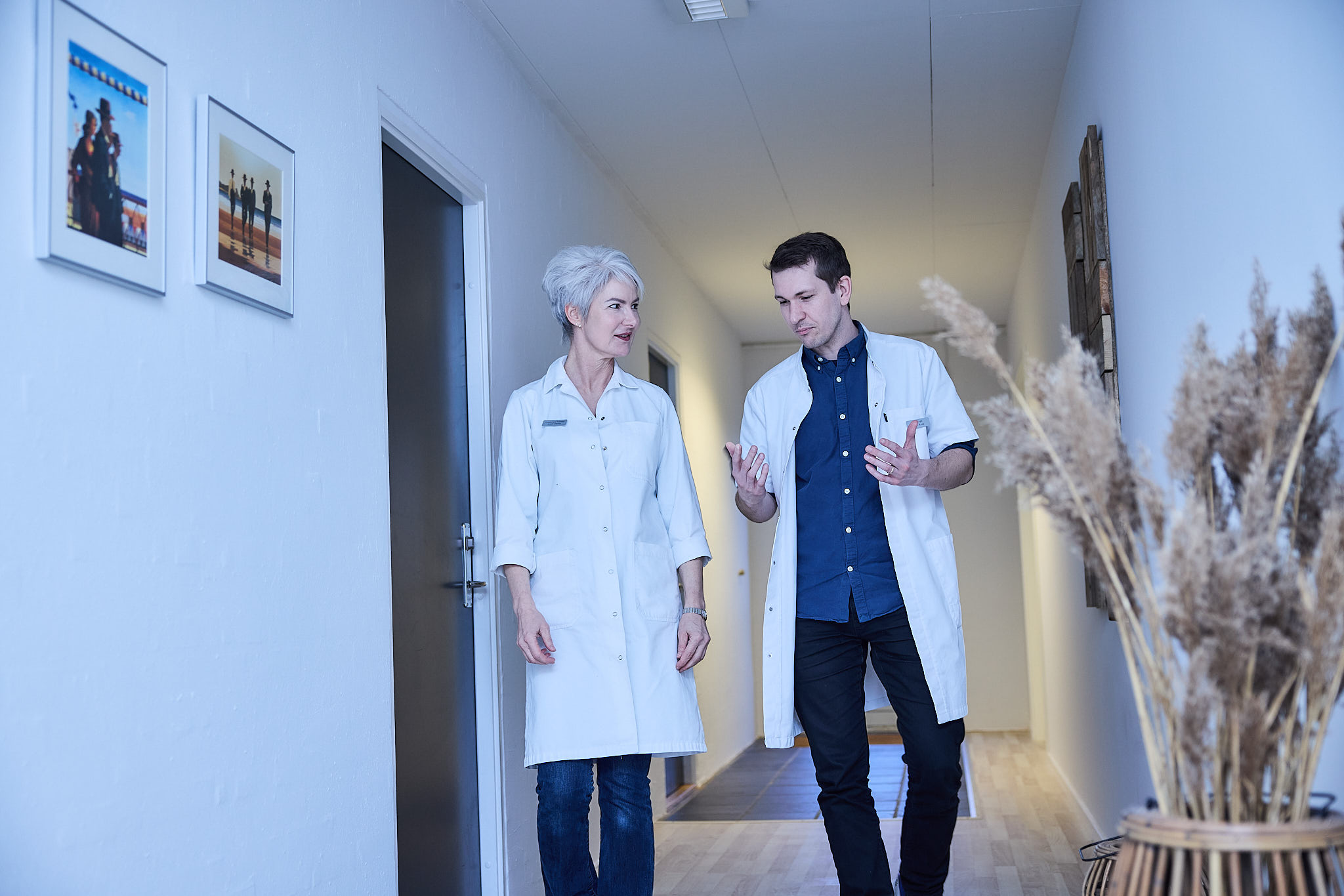 To personer i hvide labcoats går ned ad en korridor, en mand og en kvinde, der taler sammen.